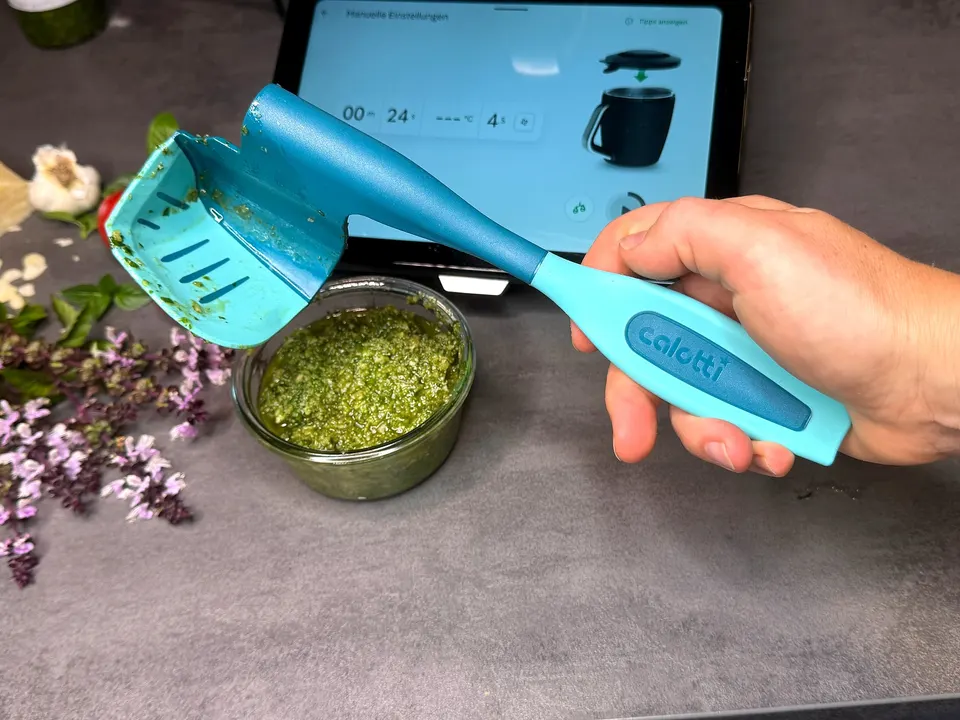 Calotti® Drehkellenspatel SILIKON+ in der Hand über einer Schale Pesto, davor Basilikumblüten